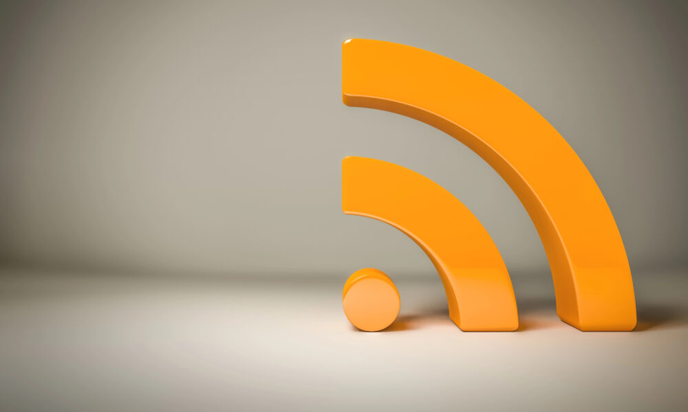 A large, three-dimensional, orange RSS symbol stands upright on a smooth, light-colored surface. The iconic RSS symbol consists of a dot at the bottom left and two curved lines radiating upward and outward to the right. The background is simple with a gradient grey.