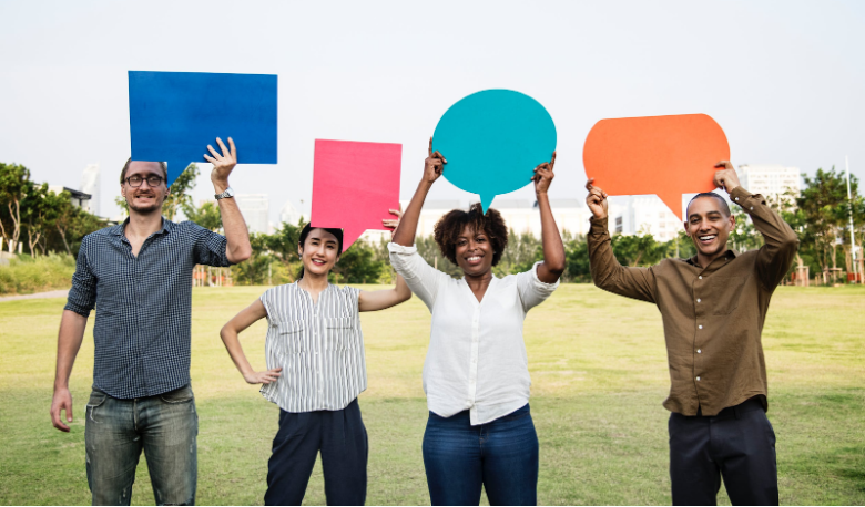 Four people standing outdoors holding up speech bubble cutouts in various bright colors. They are smiling and standing in a grassy area with trees and buildings in the background, resembling a lively discussion about social media strategy.