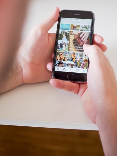 A person holding a smartphone in their left hand and scrolling through an Instagram feed with their right hand. The screen displays several images and posts relevant to their social media strategy. The phone is held over a white surface, and the person's thumb is about to tap a post.