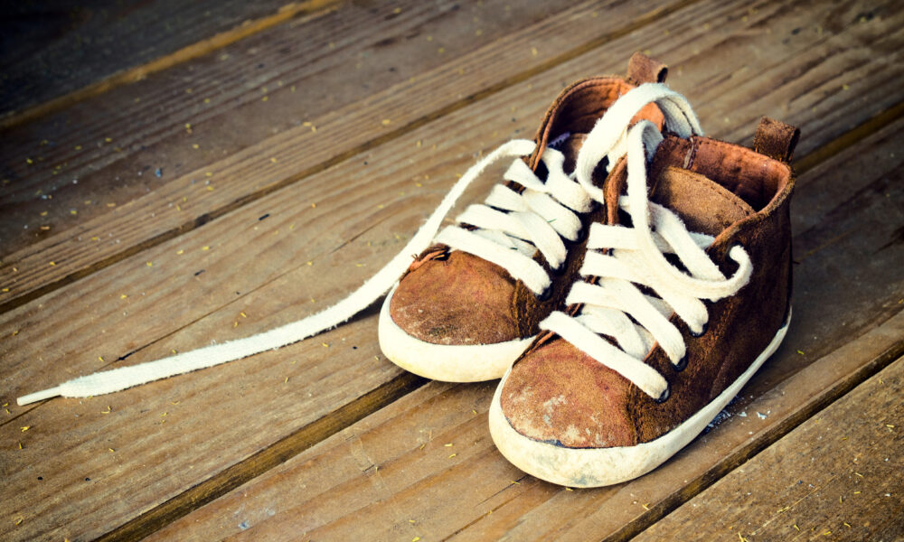 A pair of worn brown children's high-top sneakers with white laces sits on a wooden surface, perfect for any ecommerce listing. One shoelace is untied and partially trailing on the ground, adding to the charm. The shoes show signs of dirt and wear.