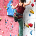A woman with braided hair is agilely rock climbing on an indoor climbing wall. She is wearing a red tank top, green pants, climbing shoes, and a harness with various climbing gear attached. The wall features colorful handholds and footholds, with sections in red and white.