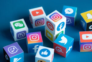 Colorful cubes with logos of various social media platforms like Facebook, Twitter, Instagram, YouTube, WhatsApp, Snapchat, WeChat, and more are scattered on a blue background. The cubes appear three-dimensional and vibrant, representing modern digital communication for the 26-year-old at home.