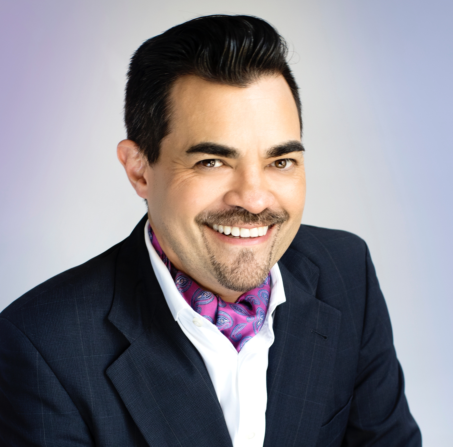 A person with short dark hair and a mustache is smiling at the camera. They are wearing a dark blazer, a white shirt, and a purple patterned scarf that adds a touch of homey elegance. The background is a gradient of light blue and purple.