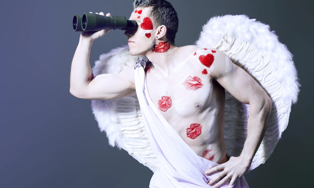 A person dressed as an angel, with white wings and wearing a draped white cloth, looks through binoculars, embodying a whimsical search intent. Their body is painted with hearts and lip prints. The background is a gradient of dark shades.