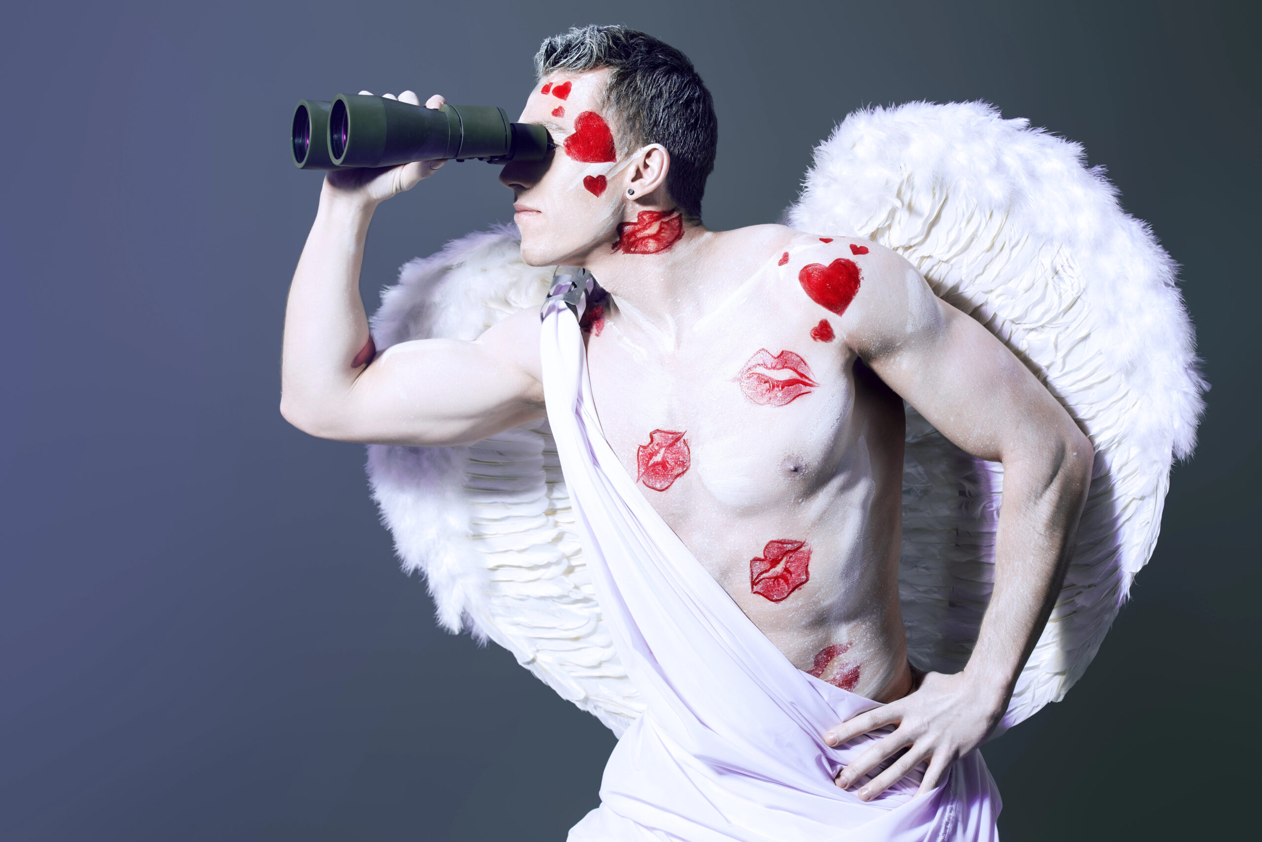 A person dressed as an angel, with white wings and wearing a draped white cloth, looks through binoculars, embodying a whimsical search intent. Their body is painted with hearts and lip prints. The background is a gradient of dark shades.
