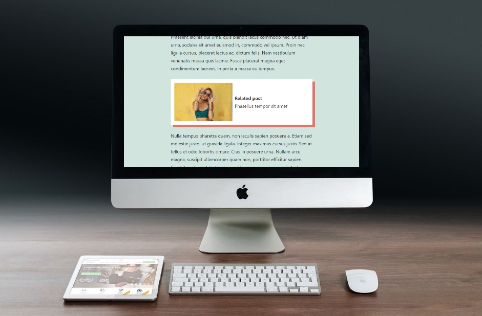 A desktop computer on a wooden desk displays a blog post with an image of a woman in a yellow top as a related post, enhancing engagement. A wireless keyboard, mouse, and a tablet showing different content are placed on the desk in front of the monitor.