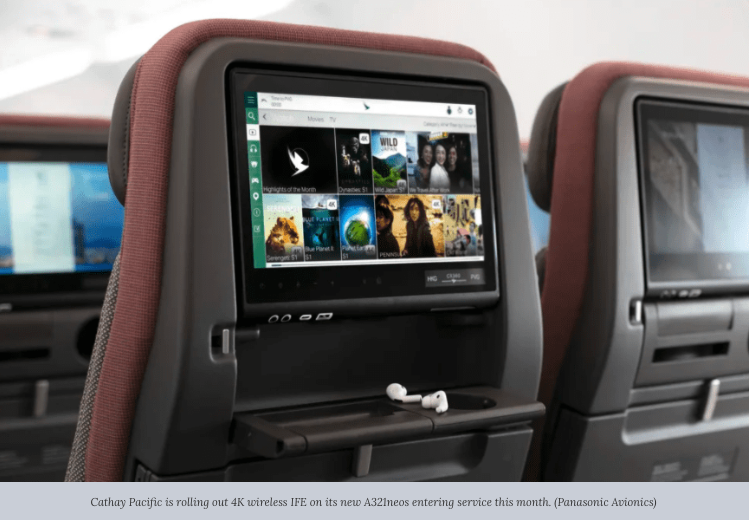 An airplane seatback features an in-flight entertainment screen displaying various movie and TV options. Meeting modern traveler expectations, the tray below holds earphones. Other seats with similar screens, part of advanced in-flight entertainment systems, can be seen in the background.