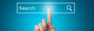 A hand is seen pointing at a virtual search bar with the word "Search" and a magnifying glass icon, all set against a blue gradient background resembling a home screen. The index finger appears to be touching the center of the search bar, emphasizing ease of use.