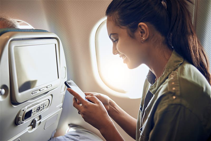 A woman sits in an airplane seat by the window, smiling as she looks at her smartphone. The seat in front features a screen and a control panel for in-flight entertainment, while sunlight streams through the window, illuminating her face. She appears relaxed and engaged with her device.