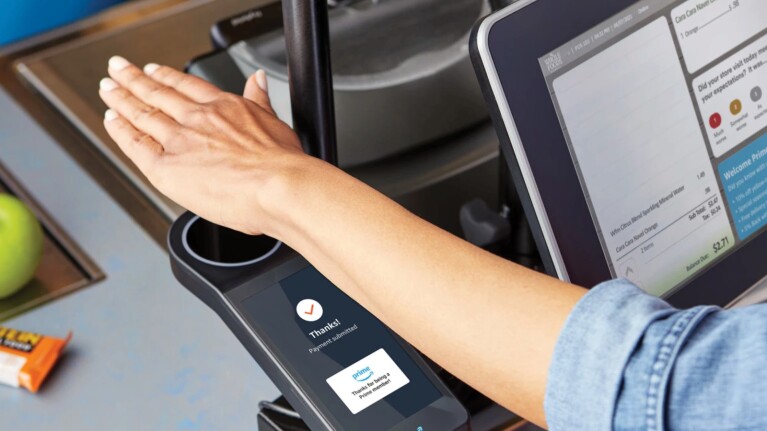 A person holds their palm over a biometric scanner at a checkout counter. The scanner screen, powered by AI, displays a message saying "Thanks!" with the Whole Foods and Amazon logos below it. A tablet or screen beside the scanner shows transaction details, showcasing the seamless integration by tech giants.